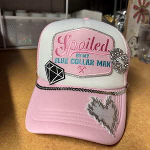 May include: A white and light pink trucker hat featuring a patch with the text Spoiled BY MY BLUE COLLAR MAN and a crossed hammer icon. The hat is decorated with a black diamond patch, a silver rhinestone lip shape, and a silver flame heart patch.
