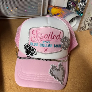 May include: A pink and white trucker hat featuring a patch with the text Spoiled BY MY BLUE COLLAR MAN and a crossed hammer icon. The hat includes a silver chain across the brim, a black diamond patch, a silver glitter heart, and a flame patch.