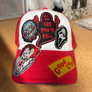 May include: A red and white trucker hat featuring embroidered patches of horror movie characters and icons. The patches include a clown, a ghost with the text SEE YOU IN HELL, a mask, and a yellow rectangular patch with the text Good Guys.