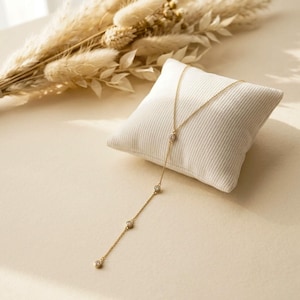 May include: A gold-toned lariat necklace featuring a delicate chain with four evenly spaced round bezel-set clear stones. The necklace is draped over a small, square, cream-colored ribbed fabric cushion, showcasing its vertical drop design.