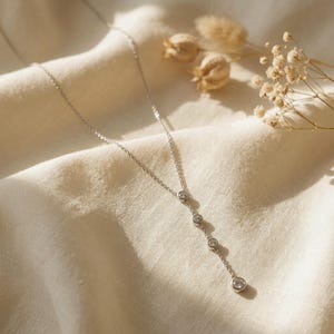 May include: A silver-toned lariat necklace featuring a delicate chain and four round, clear, bezel-set stones arranged in a vertical drop. The necklace has a polished metallic finish and a simple, refined structure that hangs in a straight line.