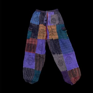Puede incluir: Pantalones de patchwork que presentan una variedad de cuadrados de tela coloridos en tonos azules, morados y marrones. Los pantalones tienen una cintura elástica con cordón y están decorados con diseños negros serigrafiados de soles y setas.
