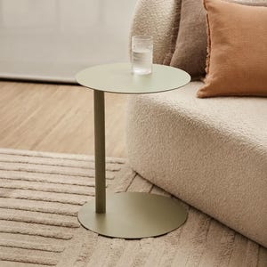May include: A sage green metal side table with a circular top, a single cylindrical support column, and a flat circular base. A clear, textured glass tumbler sits on the surface of the table, which is positioned next to a cream-colored sofa.
