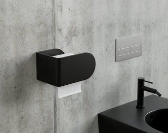 May include: A matte black wall-mounted toilet paper holder with a curved side profile, holding a roll of white toilet paper. The fixture is mounted on a grey concrete wall next to a rectangular silver flush plate and a black bathroom sink tap.