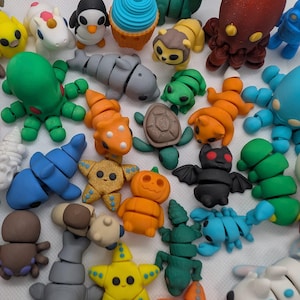 Peut inclure: Une collection de petites figurines articulées en plastique de formes et de couleurs variées, incluant des animaux comme un pingouin, une pieuvre, une tortue et une chauve-souris, ainsi qu'un cupcake, une étoile et une citrouille.