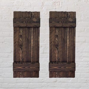 Peut inclure: Deux volets en bois brun foncé d'aspect rustique. Chaque volet est composé de planches verticales avec des bandes horizontales en haut et en bas. Le grain du bois est visible, donnant un aspect naturel et vieilli. Sur un mur de briques blanches.