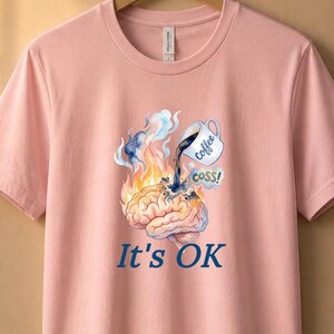 May include: A light pink short-sleeve t-shirt featuring a graphic of a brain on fire with a coffee mug pouring liquid onto it. The graphic includes the text "Coffee" on the mug, "COSS!" in a speech bubble, and the phrase "It's OK" printed below.