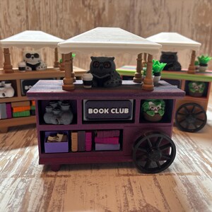 Può includere: Tre carretti in miniatura in stile legno con tetti bianchi, ognuno dotato di una piccola statuetta di gufo, una tazzina di caffè e scaffali pieni di minuscoli libri e piante in vaso. Il carretto viola centrale mostra la scritta BOOK CLUB.