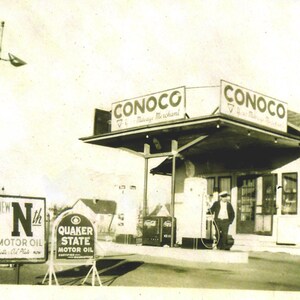 May include: A black and white photo of a gas station with a Conoco sign. The gas station has a canopy over the pumps and a sign that says "Quaker State Motor Oil".