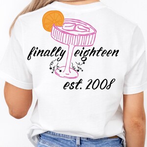 Könnte beinhalten: Ein weißes T-Shirt mit einer rosa Strichzeichnung eines Cocktailglases mit einer Orangenscheibengarnitur. Unter dem Glas steht der Text finally eighteen in schwarzer Schreibschrift, gefolgt von est. 2008 in schwarzer Schrift.