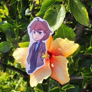 May include: A cartoon illustration of a person wearing a brown and blue patterned jacket, blue shirt, and black pants. The illustration is printed on a sticker and placed on a yellow hibiscus flower.