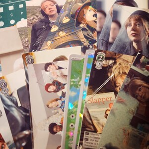 Pode incluir: Uma coleção de cartas de negociação com vários personagens e designs. Algumas cartas têm acentos holográficos. As cartas estão dispostas em uma ordem aleatória em um tapete de corte verde.