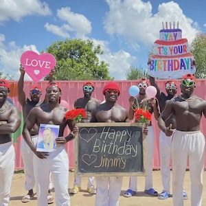 Op de afbeelding: Een groep mensen met rode petten en zonnebrillen, die rekwisieten met een verjaardagsthema vasthouden. Eén persoon houdt een bord vast met de tekst "Happy Birthday Jimmy". Anderen houden een hartvormig bord met het woord "Love" en een verjaardagstaartballon vast.