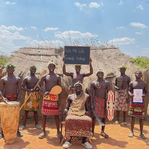 Op de afbeelding: Een groep van tien personen voor een hut met een rieten dak, gekleed in gewikkelde doeken met patronen en traditionele accessoires. Eén persoon zit op een stoel, terwijl een ander een schoolbord vasthoudt met de tekst Join the Life Guards today!