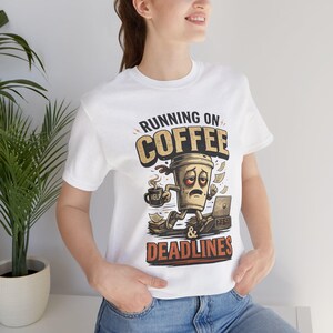 Könnte beinhalten: Ein weißes Kurzarm-T-Shirt mit einer Grafik einer müden Kaffeetasse, die einen Becher mit der Aufschrift HELP hält und vor einem Laptop sitzt, mit dem Text RUNNING ON COFFEE & DEADLINES in fetten, abgenutzten Buchstaben über und unter dem Motiv.