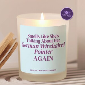 May include: A frosted glass candle with a light blue label featuring purple text that reads: Smells Like She's Talking About Her German Wirehaired Pointer AGAIN. The candle has a lit wick and a separate bamboo lid with a white silicone seal.