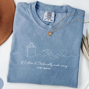 May include: A blue t-shirt featuring a white line art graphic of a shopping bag, a receipt, and a heart. The text reads, "if I return it I technically made money" and "GIRL MATH". The shirt has a Comfort Colors label showing size S.