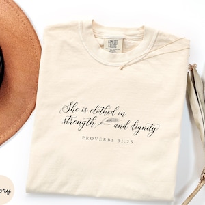 Puede incluir: Una camiseta de manga corta color marfil con texto en negro que dice "She is clothed in strength and dignity" seguido de "PROVERBS 31:25". La camiseta presenta una etiqueta de la marca Comfort Colors en el cuello y un pequeño gráfico de trigo.