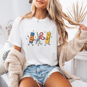 May include: A white short-sleeve t-shirt featuring a colourful graphic of a cartoon glue stick, a pair of scissors, and a ruler holding hands. The glue stick is labelled GLUE and the ruler is labelled RULER. The design includes small colourful stars.