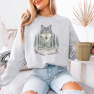 May include: A light gray crewneck sweatshirt featuring a graphic print of a wolf head above two wolves walking through a forest of birch trees. The sweatshirt has long sleeves and a ribbed neckline, cuffs, and hem, providing a relaxed fit.