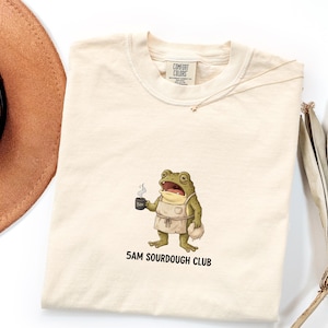 Könnte beinhalten: Ein cremefarbenes T-Shirt mit einer Grafik eines grünen Frosches, der eine Schürze trägt, eine dampfende Tasse hält und ein Brot trägt. Der Text 5AM SOURDOUGH CLUB ist unter dem Frosch gedruckt. Das T-Shirt hat ein Etikett der Marke Comfort Colors.