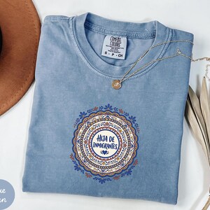 May include: A blue short-sleeved t-shirt with a round neckline and a central circular graphic featuring intricate floral patterns in orange, blue, and white. The text "HIJA DE INMIGRANTES" is printed in the center of the design with a small heart icon.