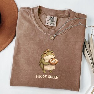 May include: A brown t-shirt featuring a graphic of a green frog wearing a small crown and an apron while holding a round loaf of bread. Below the frog, the text PROOF QUEEN is printed in a light color. The shirt has a Comfort Colors brand label.