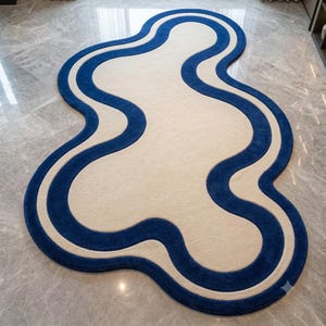 May include: An irregular, organic-shaped area rug featuring a large cream-colored center surrounded by a thick, wavy border of deep blue. The rug has a soft, textured pile and follows a fluid, non-symmetrical outline that curves and bends.