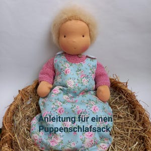 May include: A doll wearing a light blue and pink floral print sleeping bag with the text "Anleitung für einen Puppenschlafsack" on the front. The doll is sitting in a woven basket filled with straw.