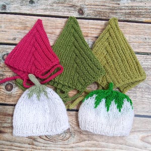 Könnte beinhalten: Eine Sammlung handgestrickter Babyhüte und -mützen in verschiedenen Farben. Die Hüte haben weiße und grüne Erdbeermotive, während die Mützen in Rosa-, Grün- und Olivtönen gehalten sind. Die Artikel sind auf einer Holzoberfläche angeordnet.