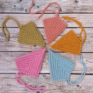 Könnte beinhalten: Fünf gestrickte Babyhauben in verschiedenen Farben: Olivgrün, Hellrosa, Senfgelb, Pink und Hellblau. Jede Haube ist dreieckig mit Rippenstruktur und Bindebändern. Die Hauben sind auf einer Holzoberfläche angeordnet.