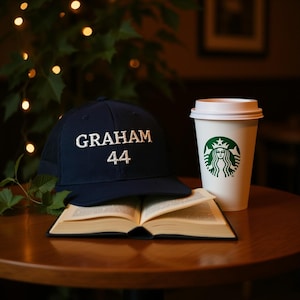 May include: A navy blue baseball cap with the name "GRAHAM" and the number "44" embroidered in white. The cap rests on an open book next to a Starbucks coffee cup. The cup features the Starbucks logo.