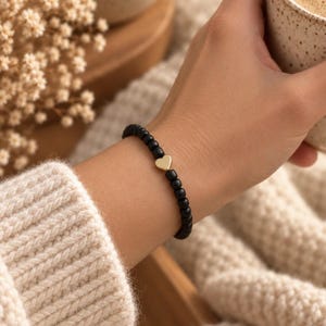 May include: A beaded bracelet featuring small, matte black spherical beads arranged in a continuous loop, accented by a single, smooth, gold-toned heart-shaped charm positioned centrally among the dark beads on the wrist.