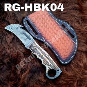 Op de afbeelding: Een mes van Damascus-staal met een gebogen lemmet, een gestructureerd gewei-handvat met drie koperen pinnen en een vingerring aan de pommel. Inclusief een bruine lederen schede met een mandvlechtpatroon en witte stiksels. Tekst leest RG-HBK04.