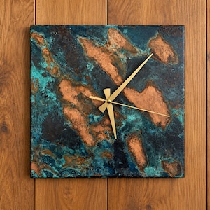 May include: A square wall clock featuring a textured, oxidized copper finish with mottled shades of teal, dark blue, and metallic copper. The clock face has three thin, gold-colored metal hands centered in the middle of the square panel.