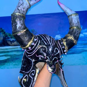 May include: A black and gold horned mask with pink and white accents. The mask has a curved shape with intricate detailing and a metallic finish. The horns are gray with pink tips. The mask is held up by a person's arm.