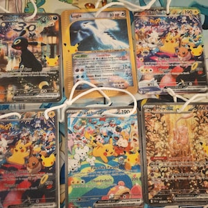 May include: Six rectangular trading cards featuring various colourful Pokémon characters, including Umbreon, Lugia, Pikachu, and Mew. Each card has a white string loop attached at the top centre, designed for hanging as a decorative ornament.
