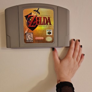 Peut inclure: Une cartouche de jeu vidéo grise pour Nintendo 64, intitulée The Legend of Zelda: Ocarina of Time. L'étiquette présente le titre du jeu en rouge, un bouclier avec une épée, une classification ESRB Everyone et le logo Nintendo 64 sur fond doré.