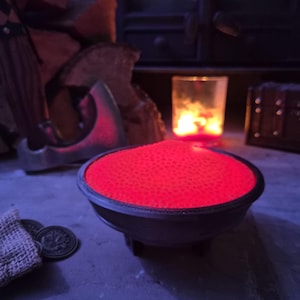 May include: A small, round, black cauldron-shaped bowl filled with a vibrant, glowing red substance that has a textured, bubbly surface. The bowl sits on three short legs and emits a bright red light, contrasting with the dark surroundings.