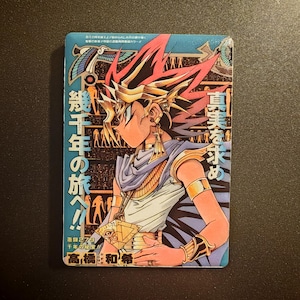 May include: A rectangular trading card featuring a colorful illustration of a character with spiky hair, wearing ancient Egyptian-style jewelry and a gold pyramid pendant. The card includes Japanese text, hieroglyphic patterns, and a blue border.
