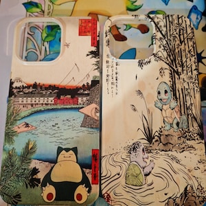 Peut inclure: Deux coques de téléphone rectangulaires ornées d'illustrations de style estampe japonaise. La coque de gauche représente un personnage Snorlax près d'un lac et d'une montagne. Celle de droite montre un Squirtle près de bambous avec du texte japonais.