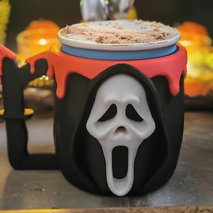 May include: A black mug featuring a white Ghostface mask design on the front, with a red drip pattern around the top rim and a black handle shaped like a knife. The mug is filled with a light-coloured beverage topped with brown sprinkles.