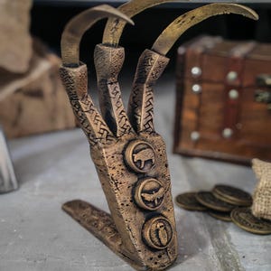 May include: A bronze-colored claw-shaped stand featuring three long, curved blades extending from a textured base. The base is decorated with three circular medallions, each displaying a different embossed animal silhouette, including a pig, a bird, and a small creature.