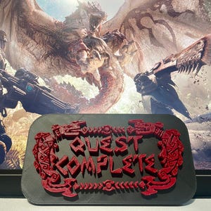 May include: A black rectangular plaque featuring the raised red text QUEST COMPLETE, surrounded by a decorative red border with intricate tribal-style patterns. The plaque has a textured finish and is displayed against a video game background.