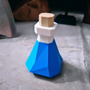 May include: A geometric potion bottle featuring a faceted blue base, a white neck section, and a tan octagonal stopper. The object has a distinct low-poly design with sharp edges and flat surfaces, creating a stylized representation of a flask.