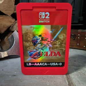 Könnte beinhalten: Eine rote, rechteckige Nintendo Switch-Spielehülle mit einem holografischen Cover für The Legend of Zelda: Ocarina of Time. Oben ist das Nintendo Switch-Logo zu sehen, unten steht der Text LB-AAACA-USA-0 auf einem schwarzen Balken.