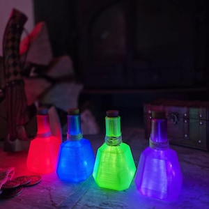 Può includere: Quattro bottiglie in vetro sfaccettato traslucido con tappi di sughero, ciascuna avvolta da uno spago attorno al collo. Le bottiglie sono illuminate dall'interno, brillando in tonalità vivaci di rosso, blu, verde neon e viola intenso.