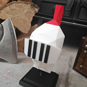 Op de afbeelding: Een wit ridderhelm-figuurtje in low-poly stijl met een felrode kam aan de bovenkant en een zwart vizier met vier verticale sleuven. De helm is gemonteerd op een vierkante zwarte voet en heeft een geometrisch ontwerp met scherpe, hoekige facetten.