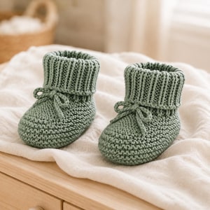 Puede incluir: Un par de patucos de punto para bebé en color verde salvia, con textura de punto bobo en el pie, puño acanalado en el tobillo y un pequeño lazo decorativo atado en la parte delantera de cada patuco, hechos de material de hilo suave.