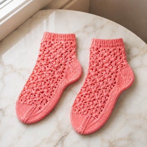 May include: A pair of coral-colored ankle socks featuring an intricate, openwork lace knit pattern throughout the body. The socks have a ribbed cuff at the top, a solid knit heel, and a solid knit toe section with a vertical seam detail at the front.